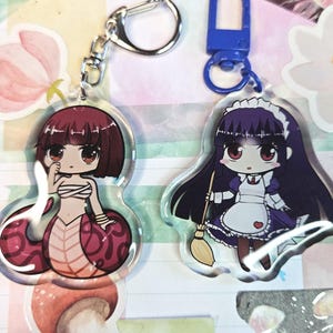 May include: Two acrylic keychains featuring anime-style characters. One depicts a snake-like figure with a red and pink design, and the other shows a maid holding a broom. Both keychains have silver and blue hardware.