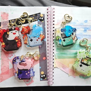 Puede incluir: Cuatro llaveros coloridos con un diseño de Game Boy, diferentes personajes y detalles florales. Los llaveros están en una libreta con el texto "Party all night" y "Sleep all day".