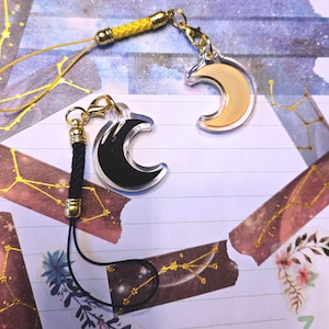 以下が含まれることがあります： 黒と金の携帯電話のチャームで、三日月型のチャームが2つ付いています。三日月型のチャームの1つは黒で、もう1つは金です。