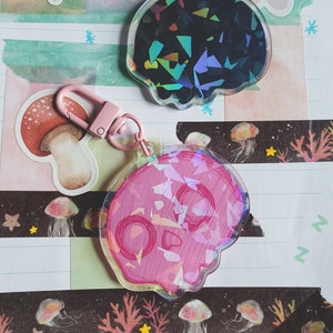 Puede incluir: Dos llaveros con forma de calavera iridiscente, uno negro y otro rosa, con un cierre de metal plateado.