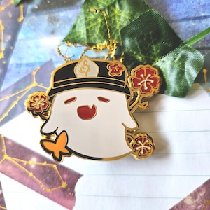 Peut inclure: Épinglette en émail doré représentant un fantôme blanc portant un chapeau noir et blanc avec un motif de feuille d'or. Le fantôme tient un petit papillon orange et est entouré de fleurs rouges et dorées.