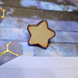 May include: A gold-colored star-shaped pin with a light yellow center and a pink outline. The pin is on a blue and white background.