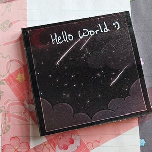 Puede incluir: Una nota adhesiva en blanco y negro con el texto "Hello World" escrito en blanco. El fondo de la nota es un cielo nocturno oscuro con estrellas y estrellas fugaces. La nota está rodeada de cinta washi rosa y blanca con diseños florales y de dibujos animados.