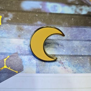 May include: A gold-colored crescent moon pin with a yellow enamel fill. The pin is on a blue and white background.