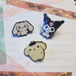 May include: Three enamel pins featuring cartoon characters. One pin is a white dog-like character with blue eyes, another is a black and white character with a skull, and the third is a tan dog-like character with a black hat.