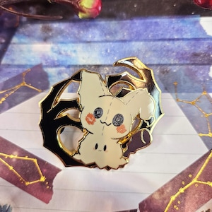 K&ouml;nnte beinhalten: Anstecknadel mit einer Cartoonfigur mit cremefarbenem K&ouml;rper, schwarzen Augen und rosigen Wangen. Die Anstecknadel ist in einen schwarzen und goldenen Rahmen gefasst. Der Hintergrund ist eine Mischung aus Lila und Blau mit dekorativen Elementen.