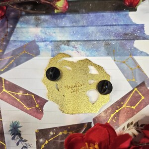 K&ouml;nnte beinhalten: Ein goldfarbener Emaille-Pin mit zwei schwarzen Nadelr&uuml;cken. Der Pin hat eine unregelm&auml;&szlig;ige Form und der Text "Masukia's Cafe" ist sichtbar. Der Hintergrund umfasst dekoratives Papier, florale Elemente und Washi-Tape mit Sternbild-Motiven.