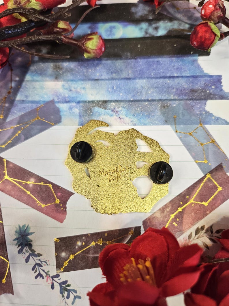 K&ouml;nnte beinhalten: Ein goldfarbener Emaille-Pin mit zwei schwarzen Nadelr&uuml;cken. Der Pin hat eine unregelm&auml;&szlig;ige Form und der Text "Masukia's Cafe" ist sichtbar. Der Hintergrund umfasst dekoratives Papier, florale Elemente und Washi-Tape mit Sternbild-Motiven.