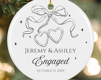 May include: White ceramic ornament with a black bow, two heart-shaped lockets, and the words "JEREMY & ASHLEY Engaged OCTOBER 15, 2025." The ornament is round and has a gold string for hanging. Black star accents surround the text.