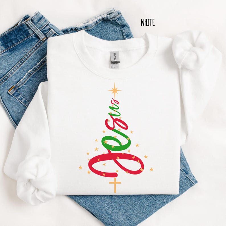 May include: White long-sleeve sweatshirt with a Christmas tree design. The tree is formed by the word "Jesus" in red and green, with a gold star at the top and a gold cross at the bottom. Gold stars are scattered around the tree.