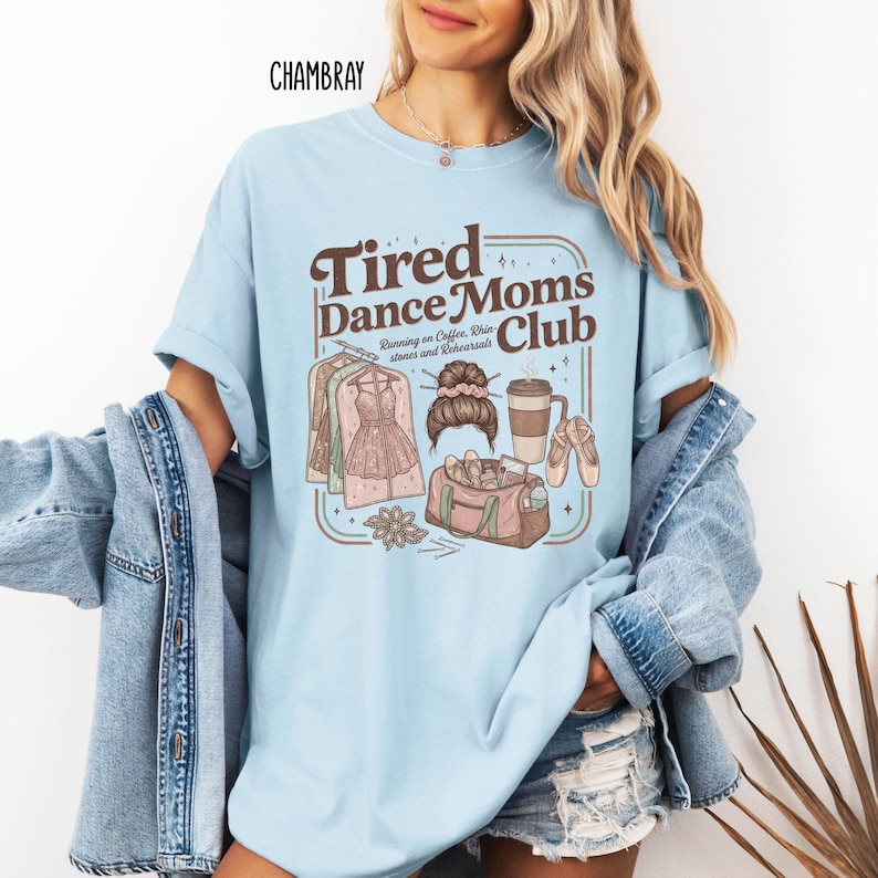 May include: Light blue t-shirt with the text "Tired Dance Moms Club" in brown lettering. The graphic includes illustrations of dance attire, a coffee cup, and a dance bag. The shirt is made of a soft material.