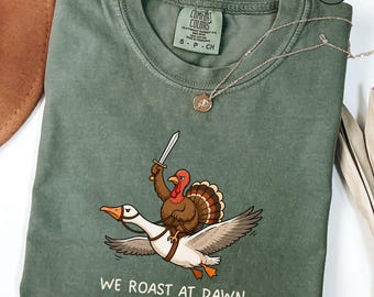 May include: A moss green crew neck t-shirt featuring a graphic of a turkey holding a sword while riding a flying goose. Below the graphic, the text reads WE ROAST AT DAWN. The shirt has a Comfort Colors brand label at the inner neckline.