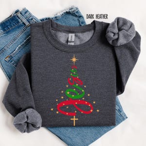 May include: Dark heather gray sweatshirt with a Christmas tree design. The tree is formed by the word "Jesus" in red and green script, with a gold star at the top and a gold cross at the bottom. Gold stars are scattered around the tree.