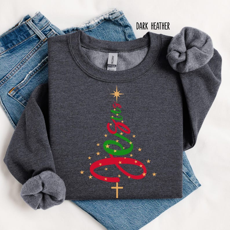 May include: Dark heather gray sweatshirt with a Christmas tree design. The tree is formed by the word "Jesus" in red and green script, with a gold star at the top and a gold cross at the bottom. Gold stars are scattered around the tree.