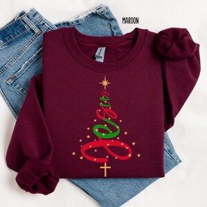 May include: Maroon sweatshirt with a Christmas tree design. The tree is formed by the word "Jesus" in red and green, with a gold cross at the bottom and gold stars. The word "Maroon" is printed at the top.