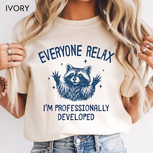 May include: An ivory t-shirt featuring a dark blue graphic of a raccoon with its paws raised, surrounded by small star icons. The text above the raccoon reads EVERYONE RELAX and the text below reads I'M PROFESSIONALLY DEVELOPED.