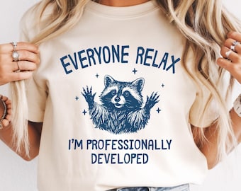 May include: An ivory t-shirt featuring a dark blue graphic of a raccoon with its paws raised, surrounded by small star icons. The text above the raccoon reads EVERYONE RELAX and the text below reads I'M PROFESSIONALLY DEVELOPED.