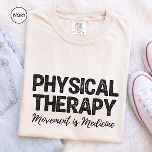 May include: An ivory t-shirt featuring the black text PHYSICAL THERAPY in a distressed, bold font, followed by the script text Movement is Medicine. The shirt includes a Comfort Colors brand label at the inner neckline.