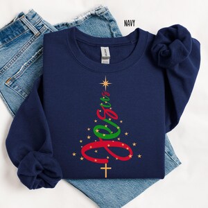 May include: Navy blue sweatshirt with a Christmas tree design. The tree is formed by the word "Jesus" in red and green script, with a gold cross at the base and gold stars. The word "NAVY" is printed in white in the top right corner.