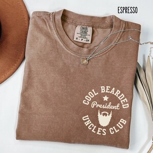 May include: Espresso-colored Comfort Colors t-shirt with a crew neck and short sleeves. The shirt features a white graphic that reads "Cool Bearded President Uncles Club" with a beard illustration. A silver necklace with a pendant is draped across the shirt.