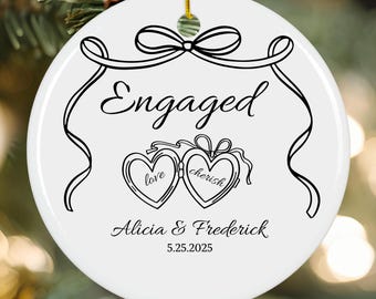 May include: White ceramic ornament with black script and line art. The word "Engaged" is written across the top. Below are two heart-shaped lockets with the words "love" and "cherish". The names "Alicia & Frederick" and the date "5.25.2025" are also included.