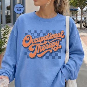 May include: A Carolina blue crewneck sweatshirt featuring a dark blue distressed checkerboard pattern on the chest. Overlaid on the pattern is the text "Occupational Therapy" written in a bold, orange, retro-style cursive font with a dark blue outline.