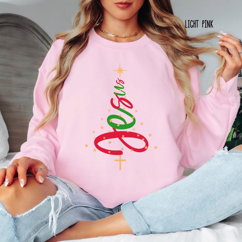 May include: Light pink sweatshirt with a Christmas tree design. The tree is formed by the word "Jesus" in red and green, with a star at the top and a cross at the bottom. Gold stars are scattered around the tree.