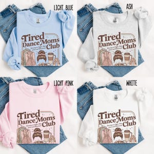 May include: Four long-sleeved sweatshirts in light blue, ash, light pink, and white. Each shirt features the text "Tired Dance Moms Club" with graphics of a dress, bun, coffee cup, and ballet shoes. The shirts are displayed on denim.
