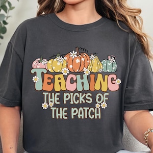 May include: Dark gray t-shirt with a retro design featuring colorful pumpkins and the text "TEACHING THE PICKS OF THE PATCH". The pumpkins are in shades of pink, yellow, orange, and green, with daisy accents.