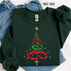 May include: Forest green sweatshirt with a Christmas tree design. The tree is formed by the word "Jesus" in red and green script, with a gold star at the top and a cross at the bottom. Gold stars are scattered around the tree.