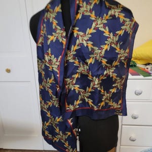 May include: A navy blue scarf featuring a repeating pattern of green holly leaves, red berries, and intertwined yellow rope accents. The scarf has a thin red border and the word OSCAR printed in gold near the bottom right corner of the fabric.