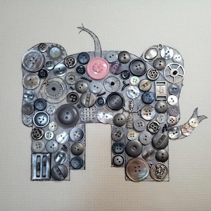 May include: An elephant design made of various gray and silver buttons, with a single pink flower-shaped button. The elephant is outlined in black on a light beige fabric. The buttons vary in size, shape, and texture, creating a unique, handcrafted look.