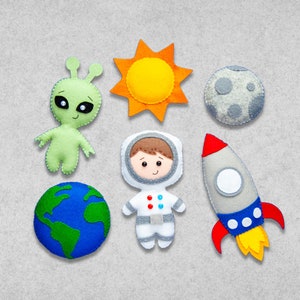 Puede incluir: Conjunto de aplicaciones de fieltro con un astronauta, un extraterrestre, un cohete, el sol, la tierra y la luna. El astronauta lleva un traje espacial blanco con un diseño azul y rojo. El extraterrestre es verde con ojos grandes. El cohete es gris con detalles rojos y azules. El sol es amarillo con rayos naranjas. La tierra es azul y verde. La luna es gris con cráteres blancos.
