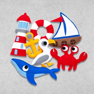 Könnte beinhalten: Ein Set aus Filzspielzeug zum Thema Nautik, bestehend aus einem rot-weißen Leuchtturm, einem gelben Anker, einer weiß-grauen Möwe, einem roten Krebs, einem blauen Wal und einem rot-weißen Rettungsring.