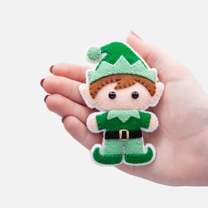 May include: A felt elf ornament with a green hat, brown hair, and a green and white outfit. The elf is holding a small red and white candy cane.