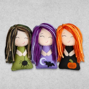 Può includere: Tre bambole di strega di feltro per Halloween con capelli e costumi di diversi colori. La bambola a sinistra ha i capelli neri e marroni e un vestito verde con un ragno. La bambola al centro ha i capelli viola e un vestito viola con una mezzaluna e un pipistrello. La bambola a destra ha i capelli arancioni e un vestito nero con una zucca.