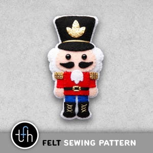 May include: A felt sewing pattern of a toy soldier nutcracker. The nutcracker is wearing a black hat with a gold emblem, a red jacket, blue trousers, and black boots. The text "FELT SEWING PATTERN" is below the image.