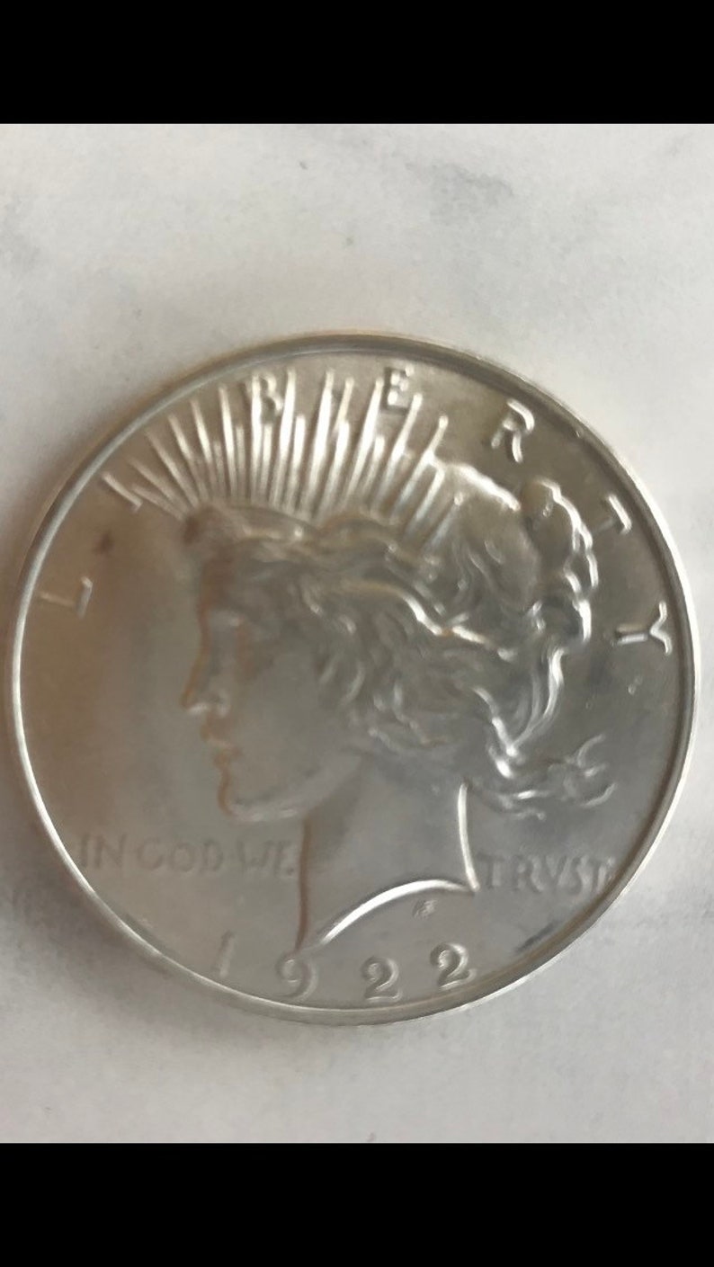 May include: A silver coin with a profile of a woman wearing a crown of rays. The coin has the inscription "IN GOD WE TRUST" and the year "1922". The word "LIBERTY" is also visible.