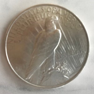 May include: A silver coin featuring an eagle perched on a branch, with the words "UNITED STATES OF AMERICA" and "E PLURIBUS UNUM" around the edge. The coin's design is detailed and the coin is in good condition.