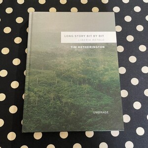 May include: A hardcover book titled "Long Story Bit by Bit Liberia Retold" by Tim Hetherington. The cover features a photograph of a lush green forest. The publisher's name, Umbrage, is at the bottom. The book is on a black and white polka dot surface.