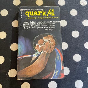 May include: A vintage paperback book titled "quark/#1" with a black cover featuring an illustration of a hand holding a needle. The cover also includes the text "a quarterly of speculative fiction" and author names.