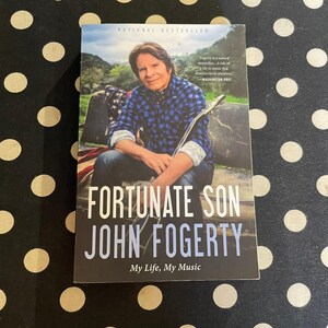 May include: A book cover for "Fortunate Son" by John Fogerty. The cover features a photo of John Fogerty holding a guitar, wearing a blue plaid shirt. The title and author's name are prominently displayed in white and blue. The book is a National Bestseller.