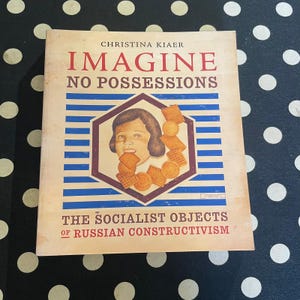 May include: A book titled Imagine No Possessions: The Socialist Objects of Russian Constructivism by Christina Kiaer. The cover features a beige background with blue horizontal stripes, a central hexagon, a child's face, and various crackers.