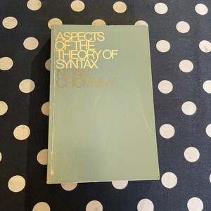 May include: A vintage book titled "Aspects of the Theory of Syntax" by Noam Chomsky. The book has a light green cover with gold lettering. The book is lying on a black and white polka dot surface.