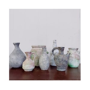 May include: A selection of antique pottery vessels in diverse shapes and sizes. The pieces display shades of grey, beige, and green, with signs of wear. The vessels are arranged on a dark brown surface against a white backdrop.