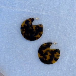 May include: A pair of brown and black tortoise shell earrings with a semi-circle shape. The earrings have a gold-tone post.
