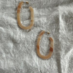 May include: A pair of oval-shaped hoop earrings made of brown and white marbled resin. The earrings have a gold-toned metal post.