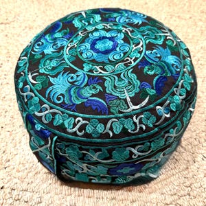 May include: Round meditation cushion with a black fabric base and blue and green embroidered floral design.