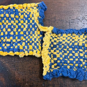 May include: Two handmade potholders. The potholders are woven in a blue and yellow checkerboard pattern. The edges are trimmed with a ruffled blue and yellow border. One potholder has a yellow loop for hanging.
