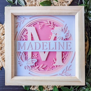 May include: A wooden frame with a white background and a pink and white layered paper design featuring the letter "M" and the name "Madeline". The design includes butterflies and floral elements.